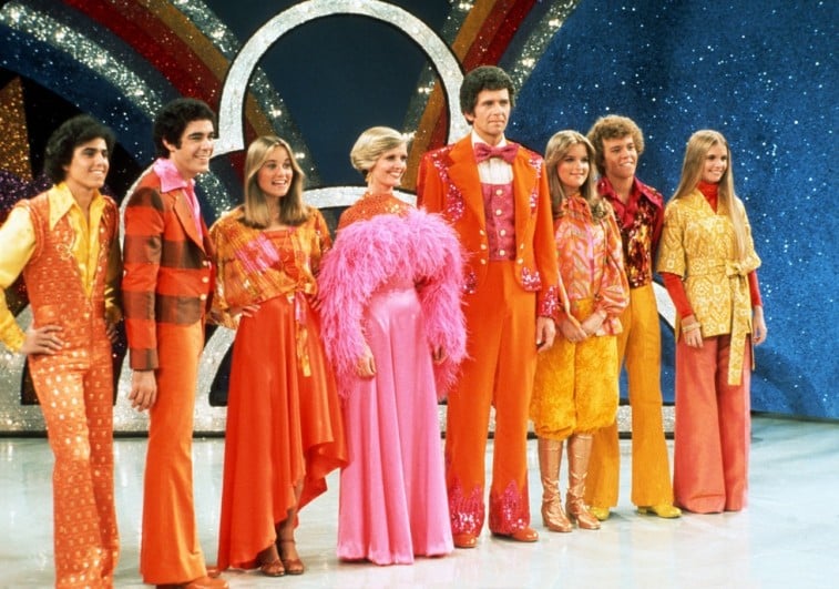 The Brady Bunch cast stand on stage in a line while wearing trajes coloridos em The Brady Bunch Hour