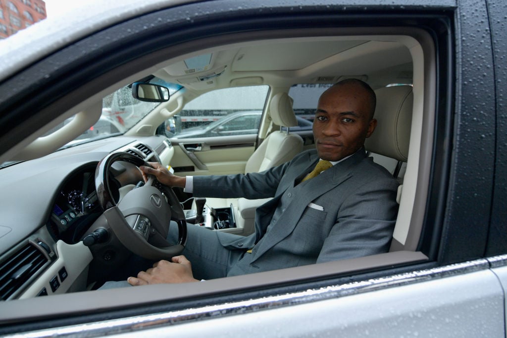 A chauffeur in a gray suit looking out the window of a car at the camera