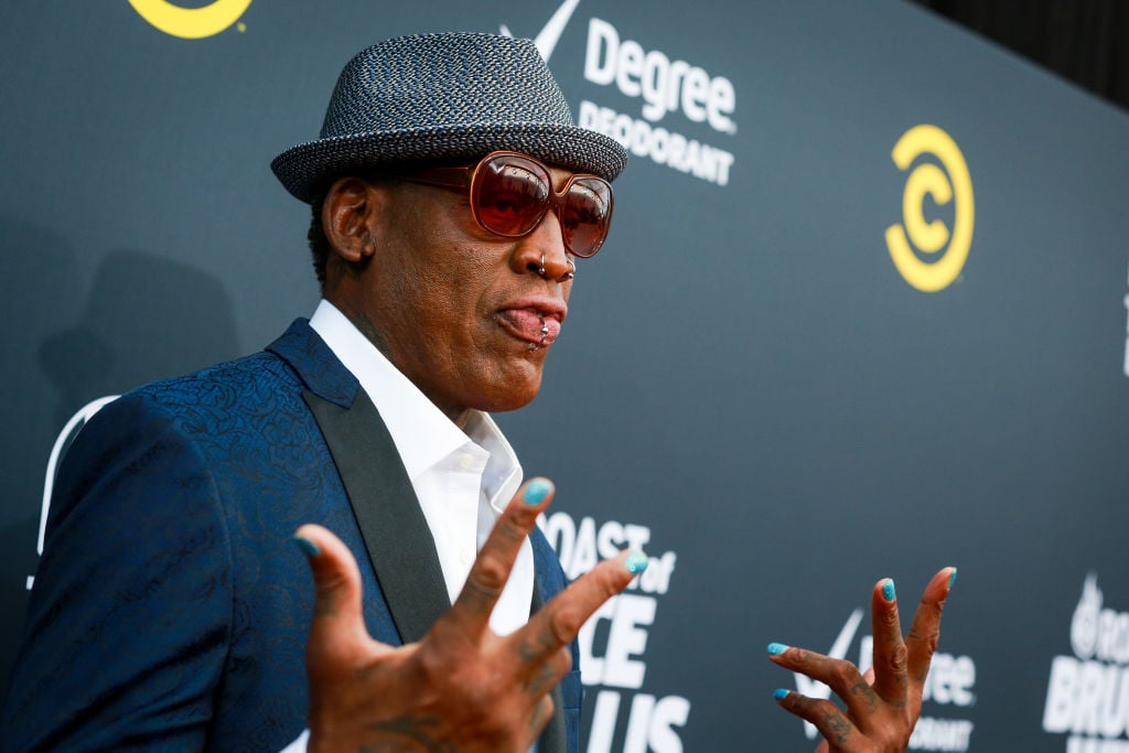 Dennis Rodman holding out his hands, smiling slightly in front of a repeating background