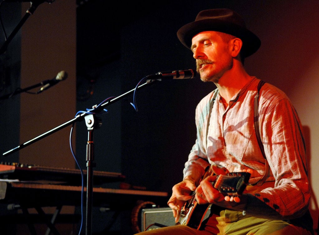 Billy Childish sitting, wearing a fedora, playing a guitar and singing into a microphone