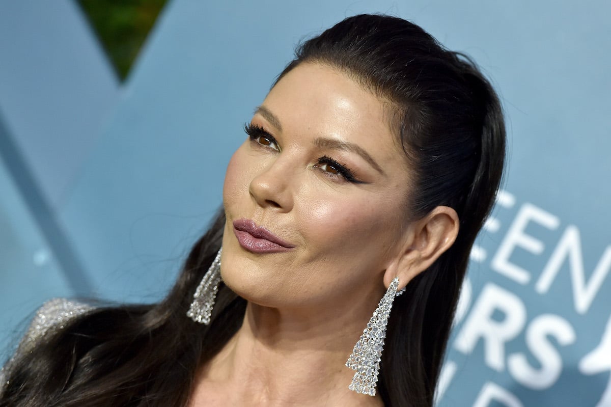 Catherine Zeta-Jones smirks with her hair half pulled back. She wears long dangling diamond earrings and stands in front of a light blue backdrop.