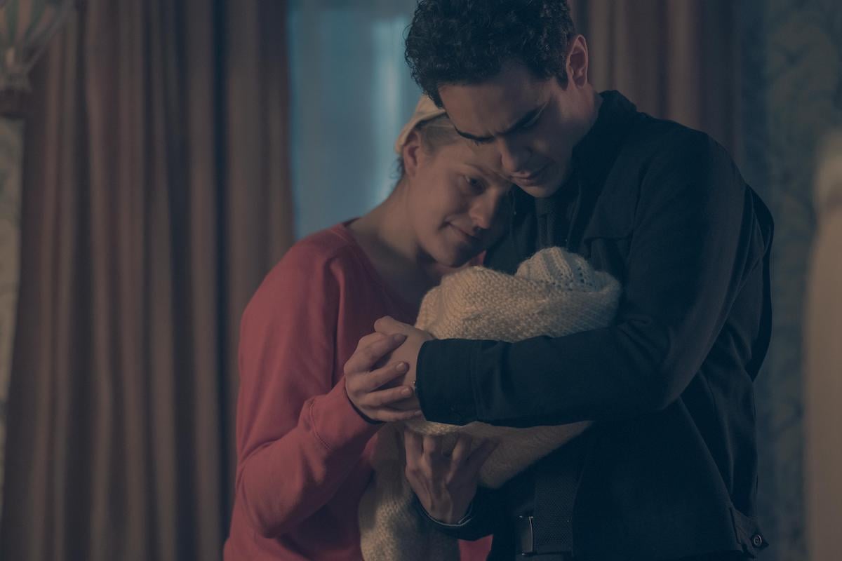 Elisabeth Moss in a red dress and white bonnet as June, Max Minghella in a black shirt and pants holding a baby wrapped in a white quilt in 'The Handmaid's Tale' Season 2