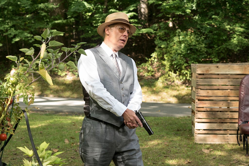 James Spader as Raymond 'Red' Reddington hides outside with his gun drawn. He's wearing a tan fedora, sunglasses, vest, nice slacks, and a white button-up shirt.