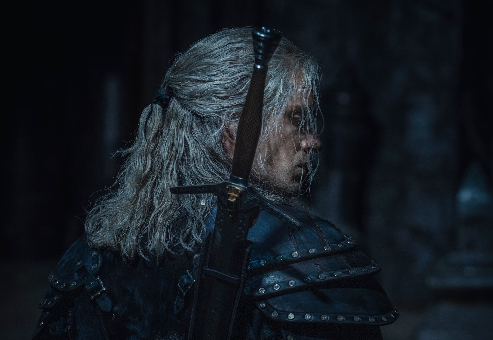 Henry Cavill as Geralt of Rivia in Netflix's 'The Witcher,' which has a prequel series ('The Witcher: Blood Origin') currently in production. Cavill wears dark armor and is facing away from the camera. His blonde hair is messy and he's looking to the side.