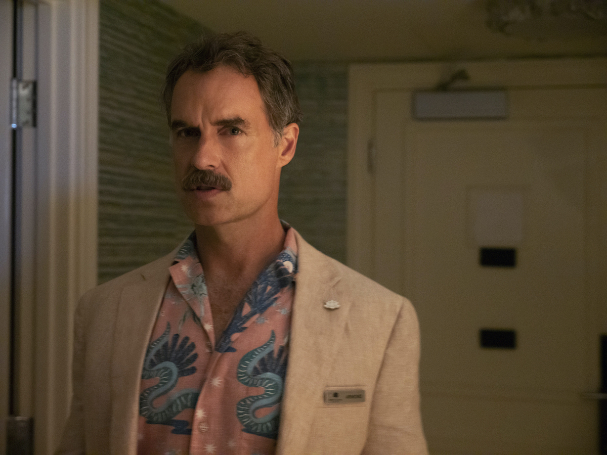 Murray Bartlett as Armond in 'The White Lotus' on HBO. The photo is from 'The White Lotus' poop scene in the season 1 finale, during which Armond sneaks into Shane Patton's room and poops in a suitcase. He stands in the entryway of a hotel suite wearing a pink button-down shirt with blue tropical print and a light beige suit jacket.