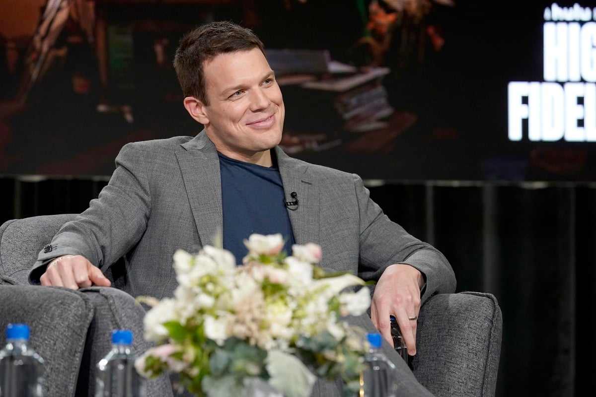 Jake Lacy in a gray suit and blue shirt in front of a bouquet of flowers.