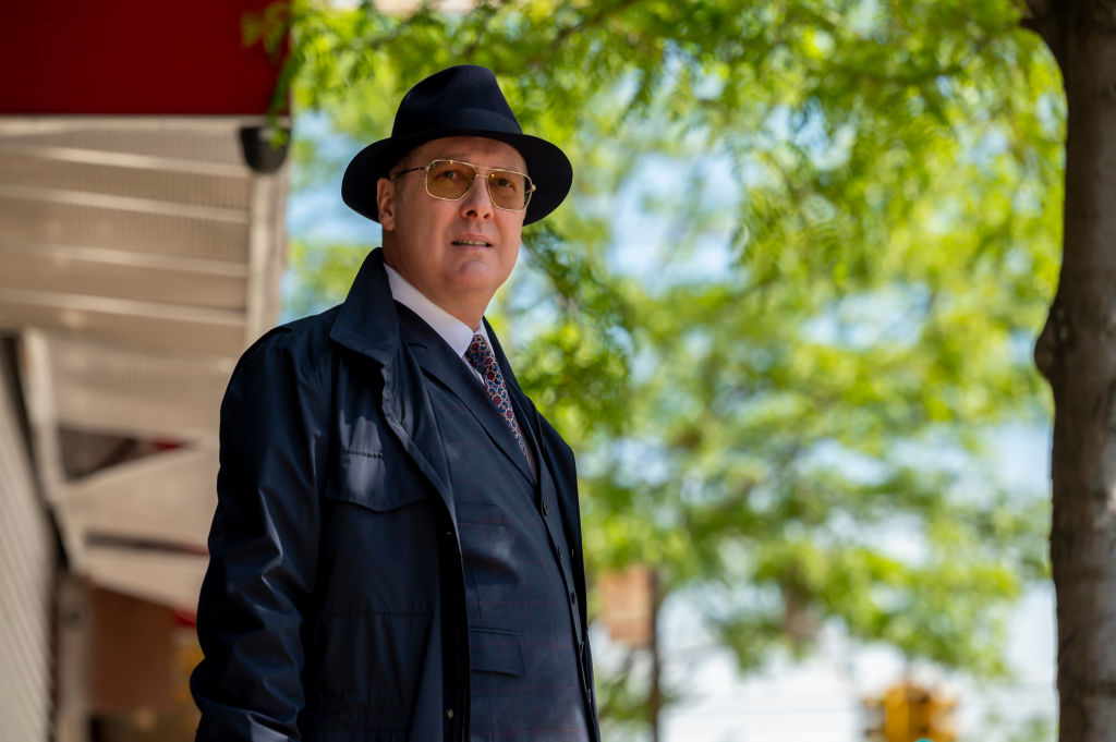 'The Blacklist' Season 9 star James Spader as Raymond 'Red' Reddington is dressed in a navy suit with matching trench coat and fedora as he peers through his sunglasses while standing outside.