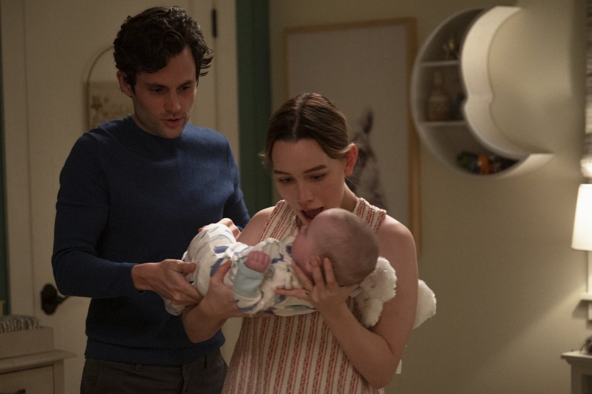 Love holds baby Henry while Joe stands behind her. Love is wearing a striped top and Joe is wearing a blue sweater.