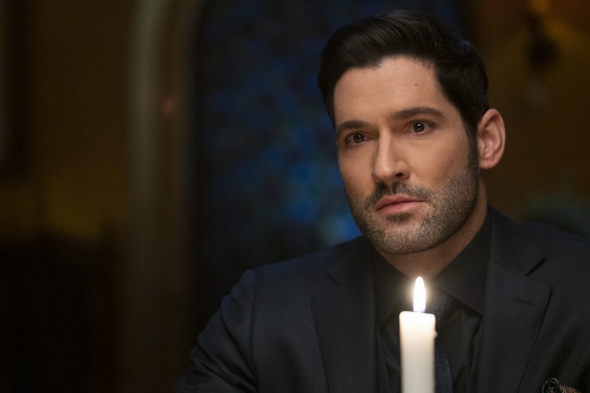 Tom Ellis as Lucifer Morningstar in the spooky series 'Lucifer.' Lucifer is sitting behind a lit candle wearing an all-black suit.