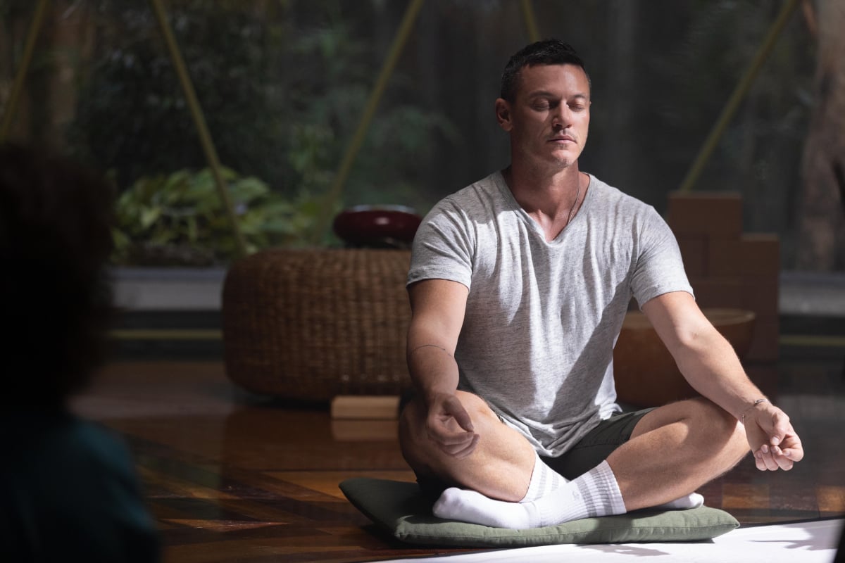 Lars sits cross-legged on a pillow with his eyes closed and meditates in 'Nine Perfect Strangers.' He is wearing a t-shirt, shorts, and long white socks.