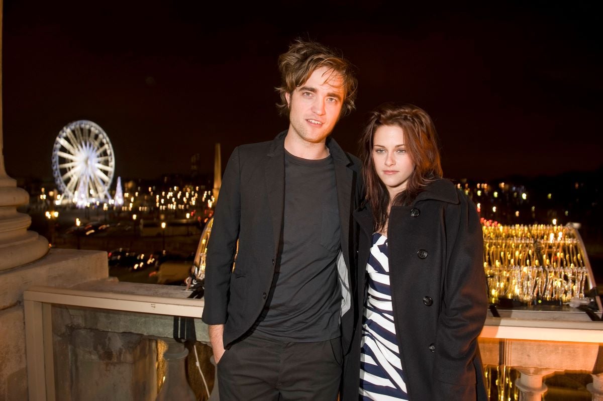 Robert Pattinson in black and Kristen Stewart in black and white, standing close with Paris in background