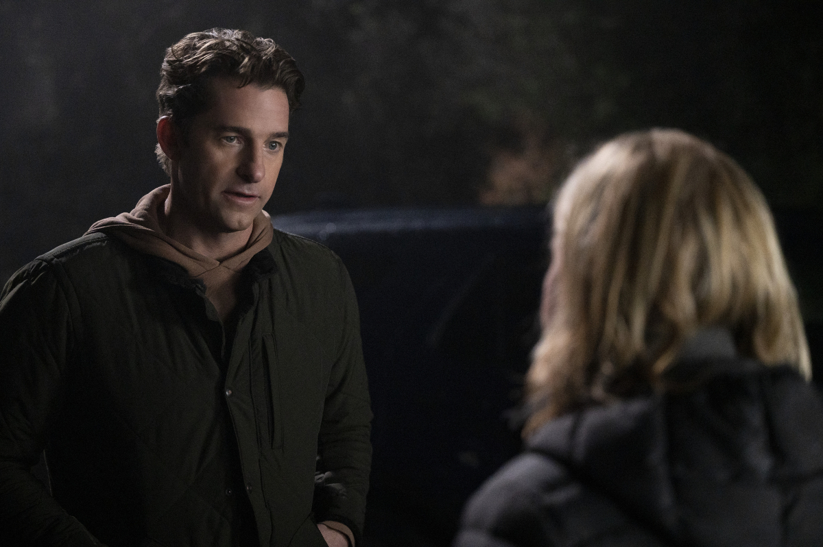 Scott Speedman as Matthew Engler talks to Romy Rosemont as Detective Falco. Matthew is wearing a jacket and Detective Falco's face cannot be seen.