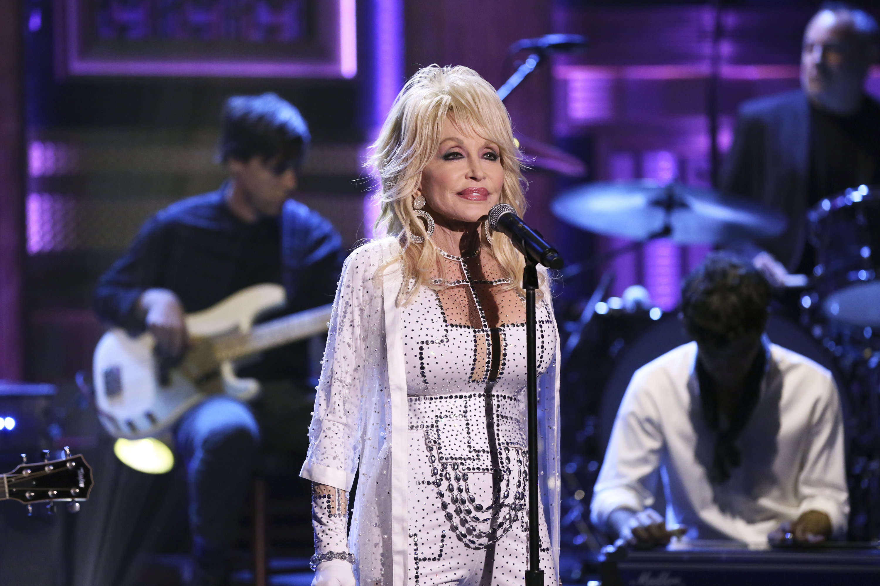 Dolly Parton wears a white dress with silver rhinestones and stands in front of a microphone.