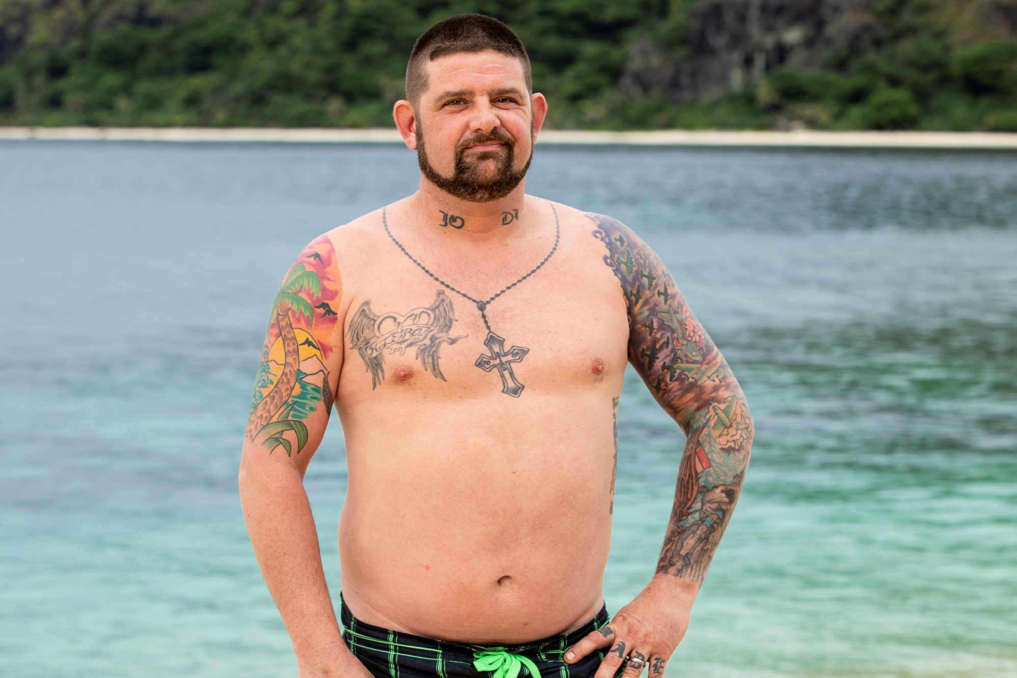 'Survivor' Season 37 contestant Pat Cusack poses shirtless on the beach, wearing green and black swim trunks. He has many tattoos on his arms and chest.