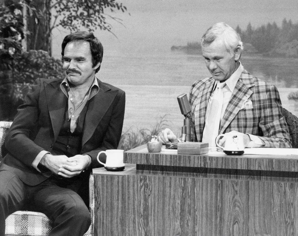 Burt Reynolds leans on the 'Tonight Show' desk next to Johnny Carson, who is wearing a plaid jacket