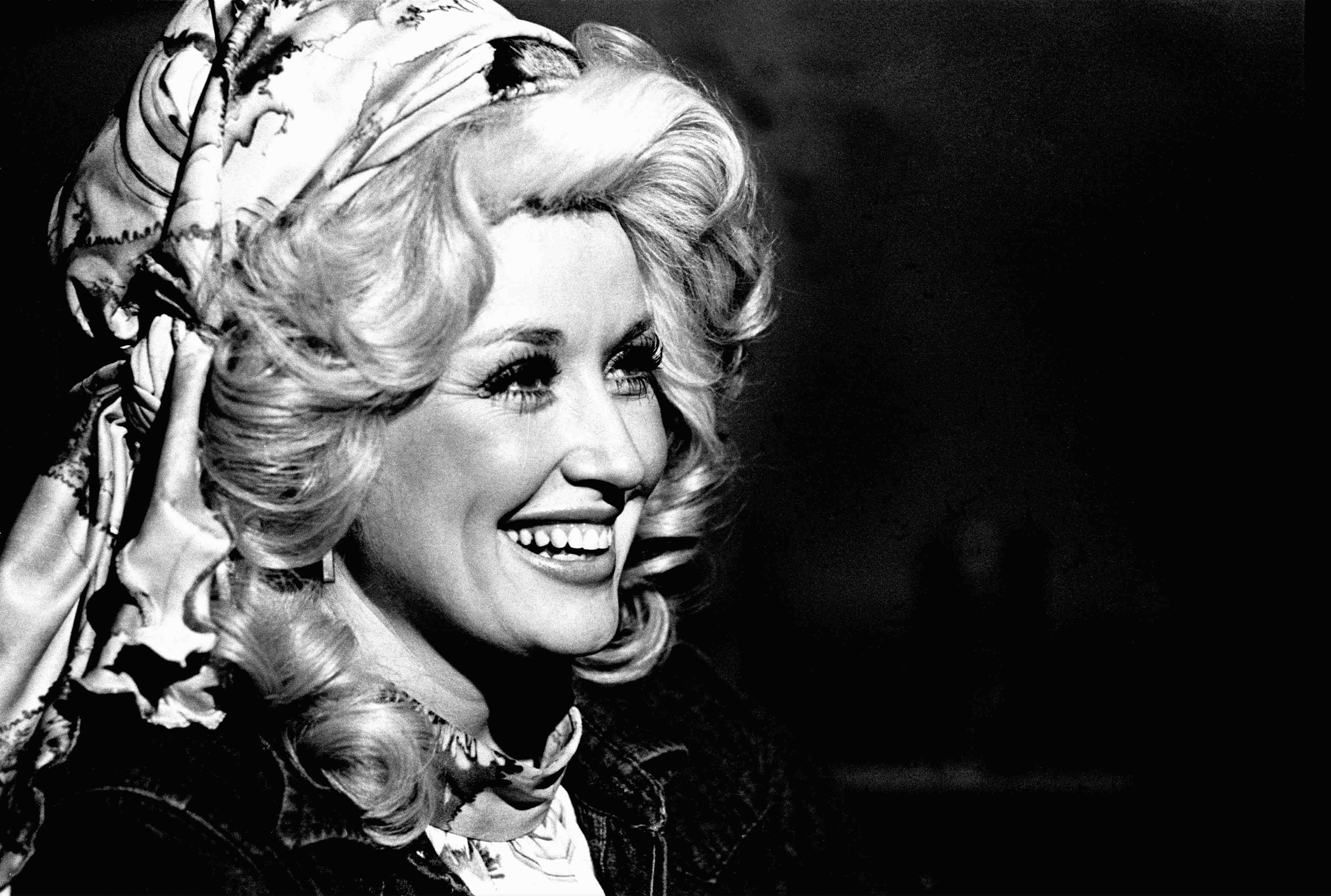 A black and white photo of Dolly Parton wearing a jacket and a scarf around her hair.