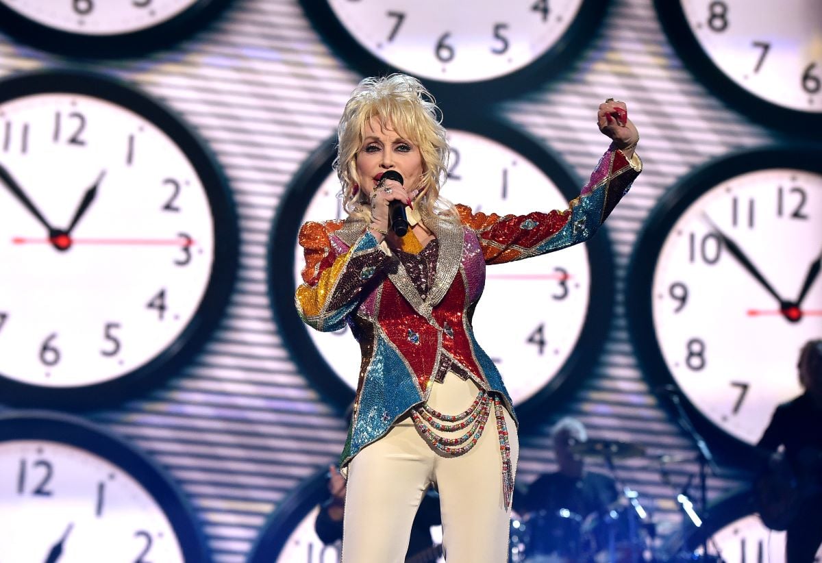 Dolly Parton in a sparkly, colorful top, holding a microphone with one hand and holding the other hand up