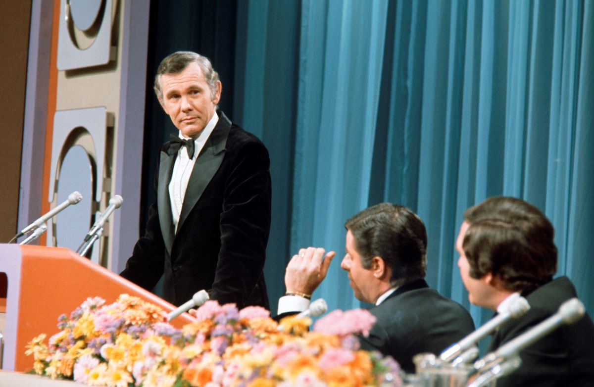 Johnny Carson in a black suit and bowtie, standing behind a podium