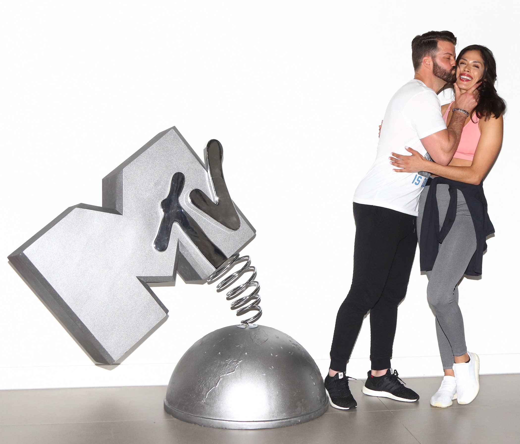 Johnny 'Bananas' Devenanzio from MTV's 'The Challenge' kissing Nany Gonzalez on the cheek while Nany laughs. They're standing next to a chrome MTV statue.