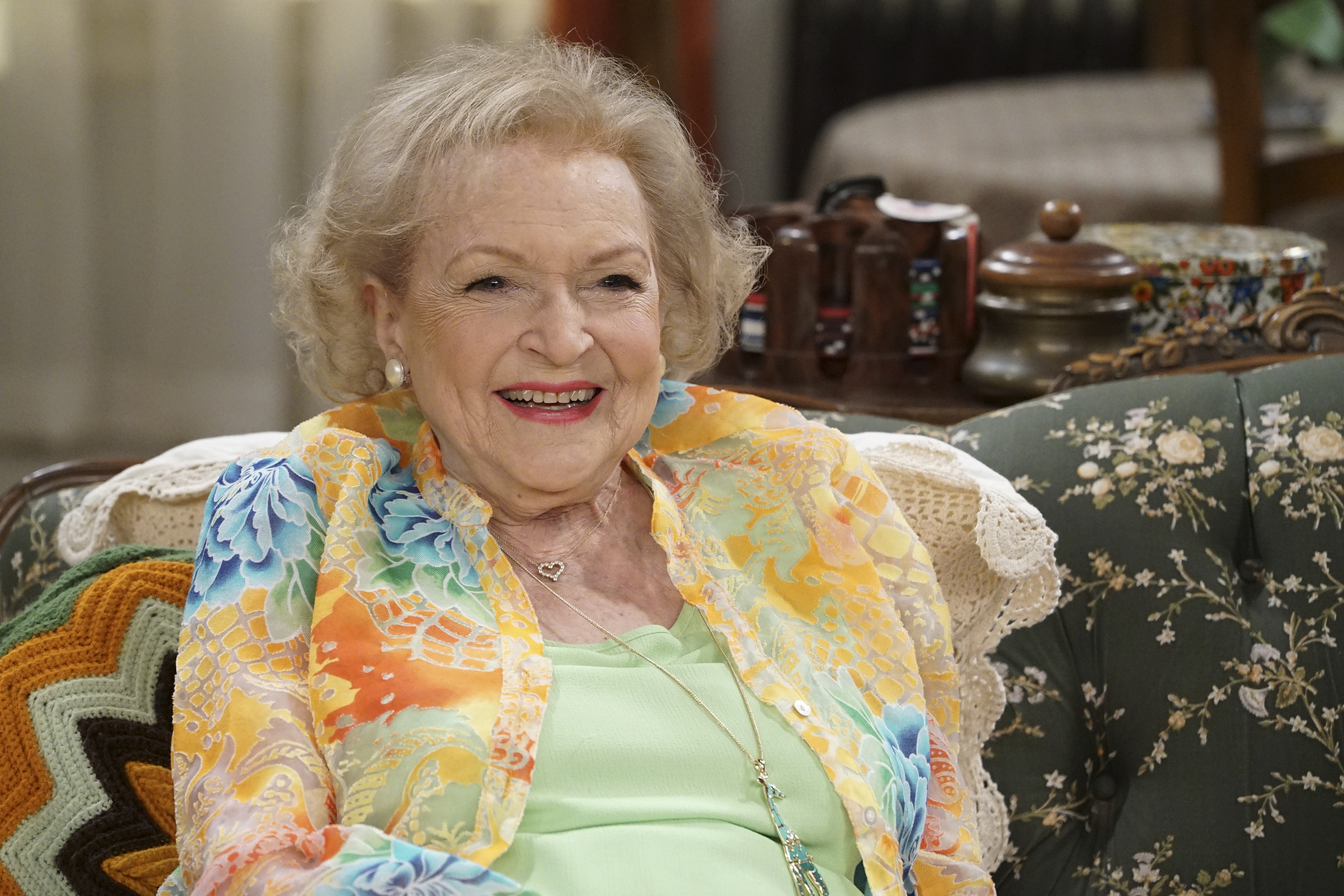 Betty White wearing a green shirt and floral blouse; sits on a couch.
