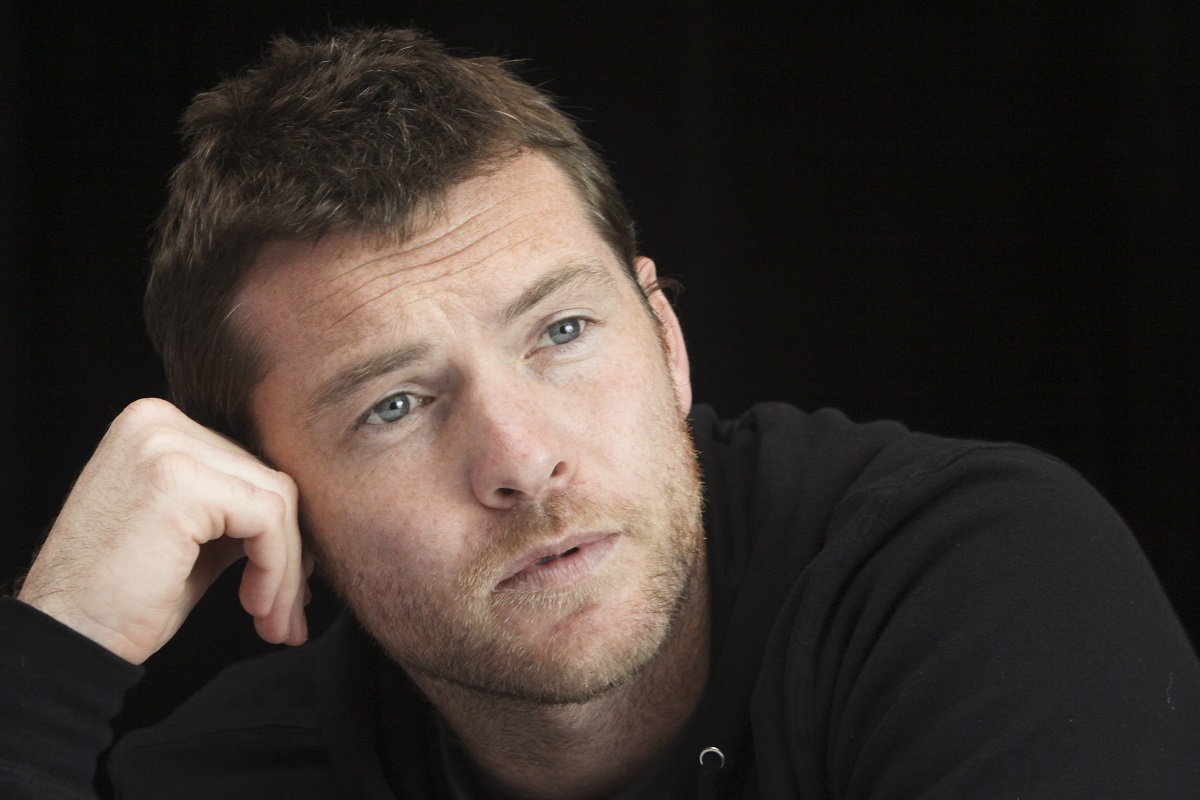 Sam Worthington sitting down while wearing a black sweater.