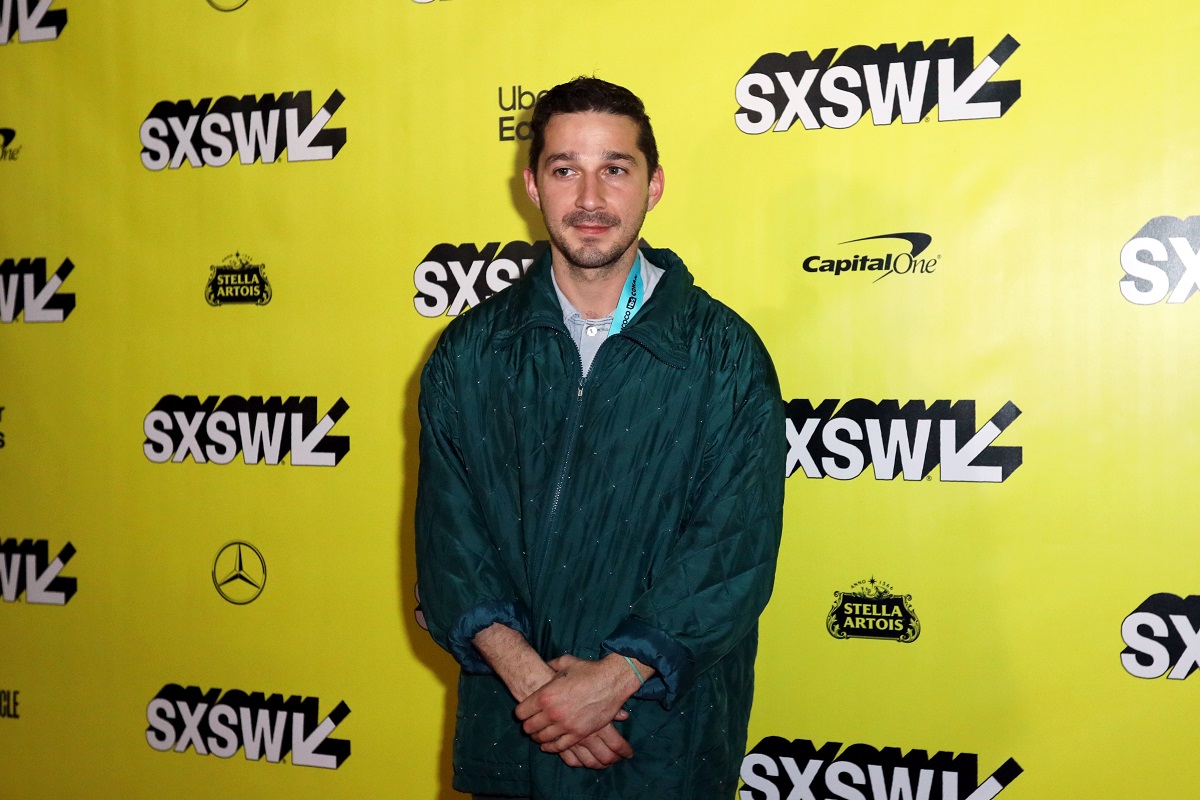 Shia LaBeouf smiling while wearing a coat.