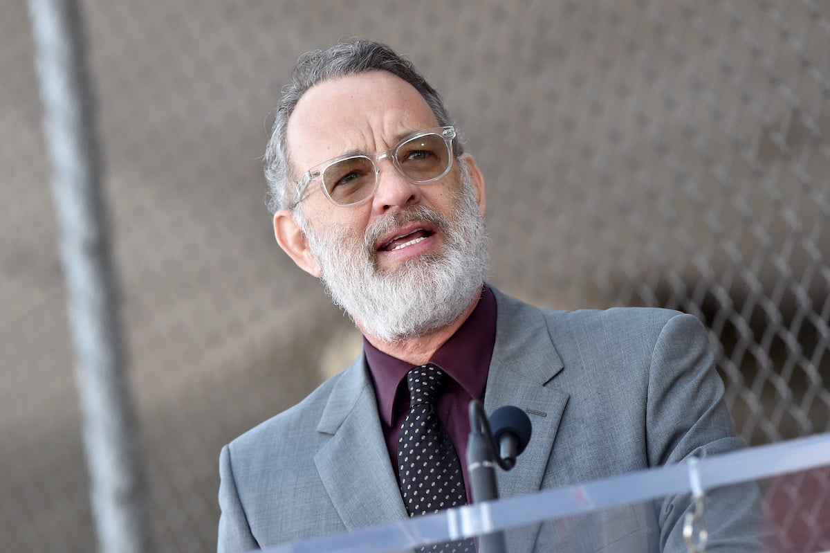 Tom Hanks wears a gray suit and glasses as he speaks at a mic