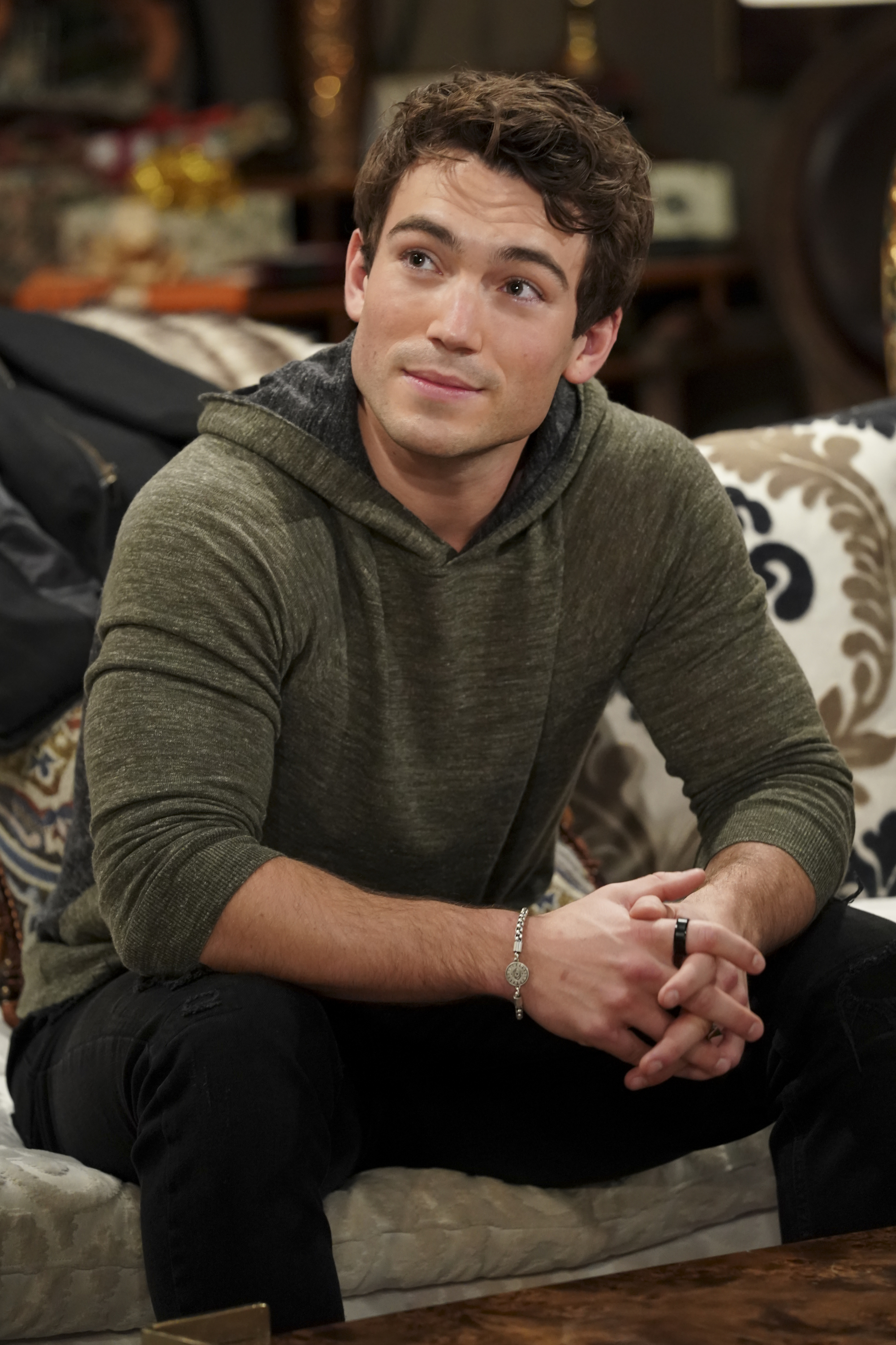 'The Young and the Restless' actor Rory Gibson wearing black pants and a grey hoodie; sits on a couch.