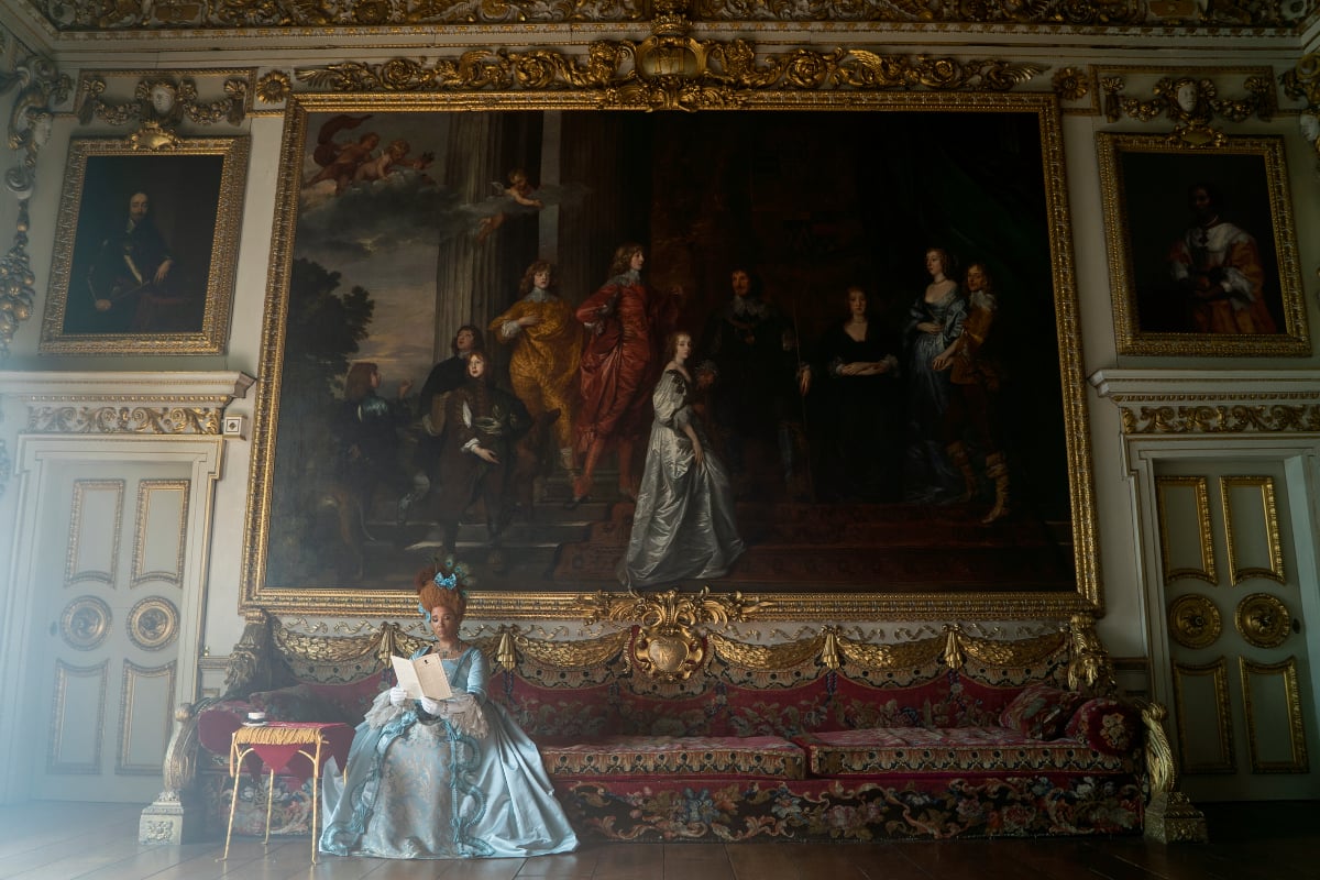 Golda Rosheuvel as Queen Charlotte in Bridgerton Season 2. Queen Charlotte reads in front of a large painting.