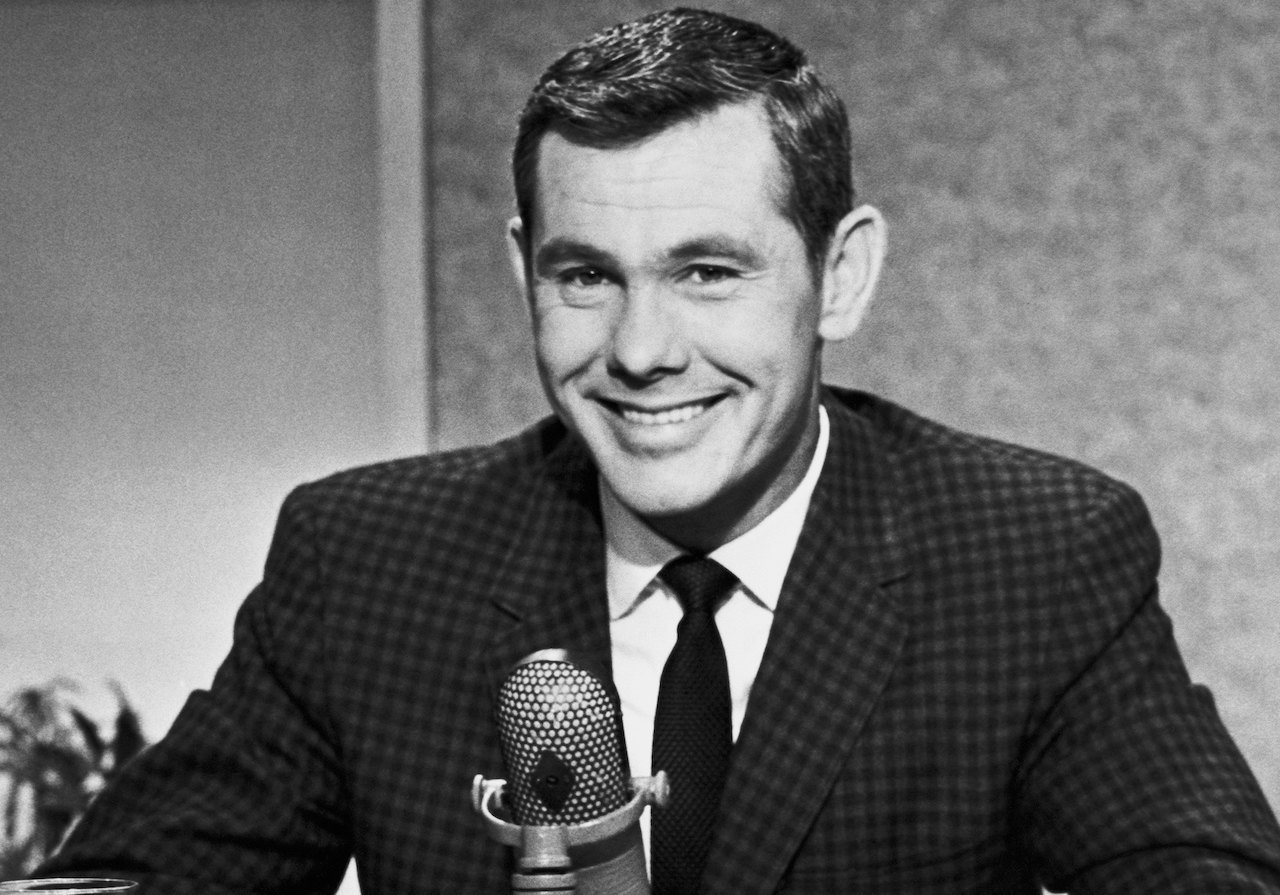 Black and white photo of Johnny Carson in a suit, seated in front of a microphone and smiling c. 1963