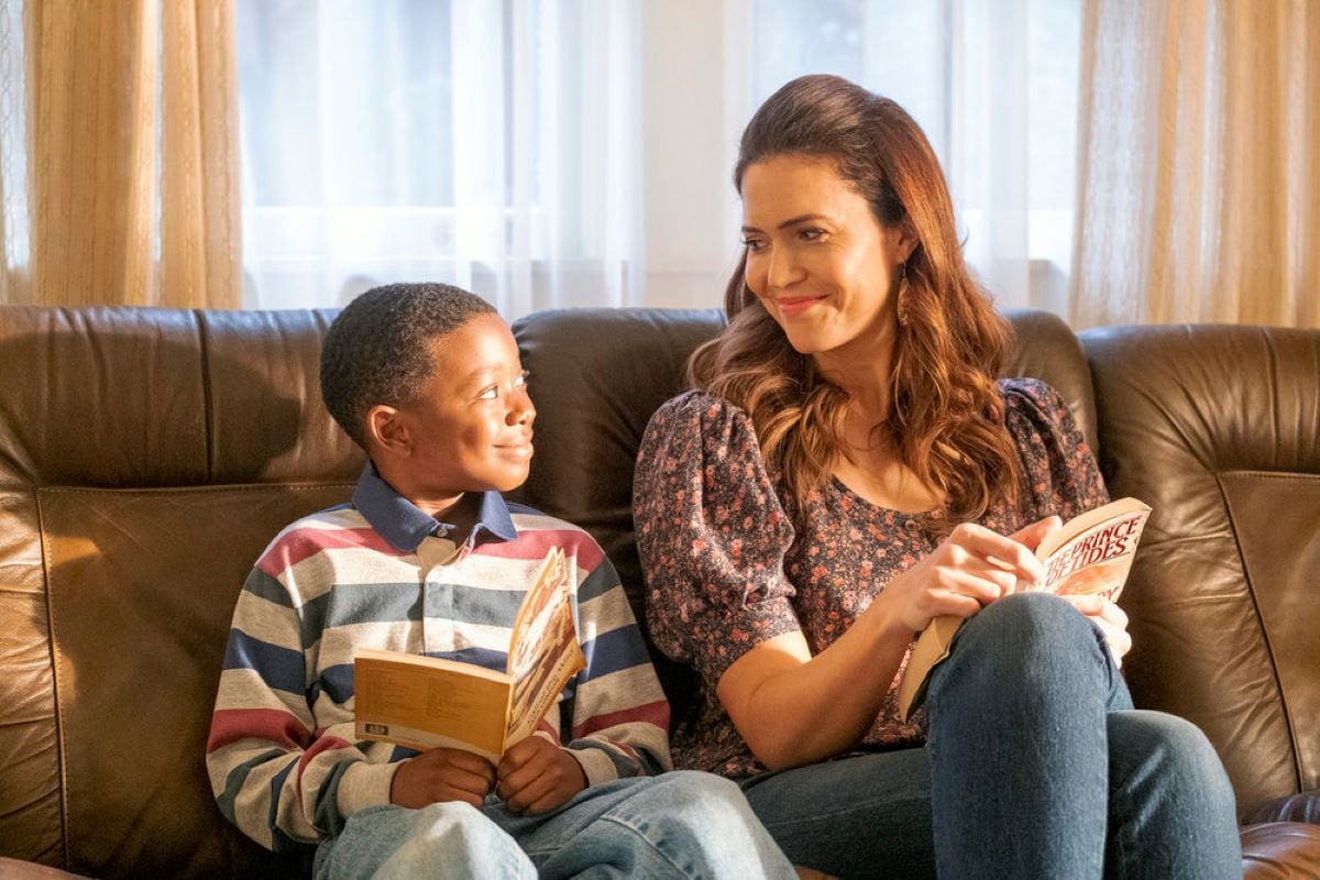 Ca’Ron Jaden Coleman and Mandy Moore share a scene as Randall and Rebecca on a couch in 'This Is Us' Season 6 Episode 10, 'Every Version of You.' Randall wears a red, blue, and gray striped long-sleeved shirt and jeans, and he holds an open book. Rebecca wears a black and pink floral shirt and jeans, and she holds an open book.