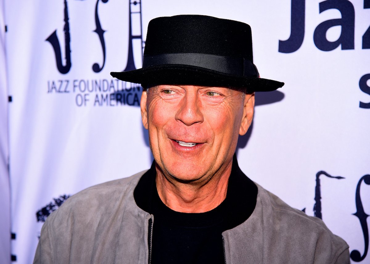 Bruce Willis wears a dark hat and smiles