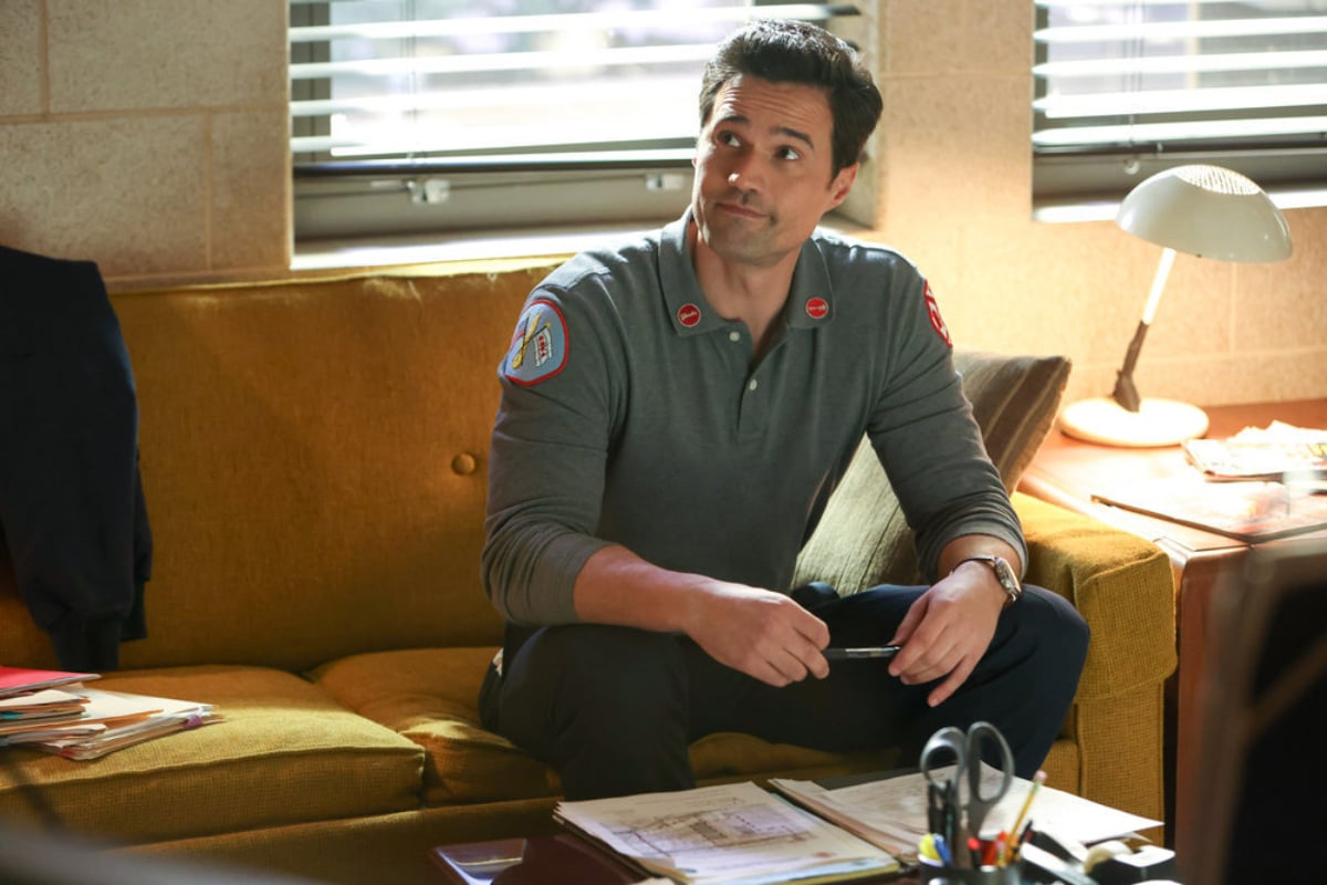 Brett Dalton as Jason Pelham in Chicago Fire Season 10. Pelham sits on the couch wearing a grey CFD shirt. 