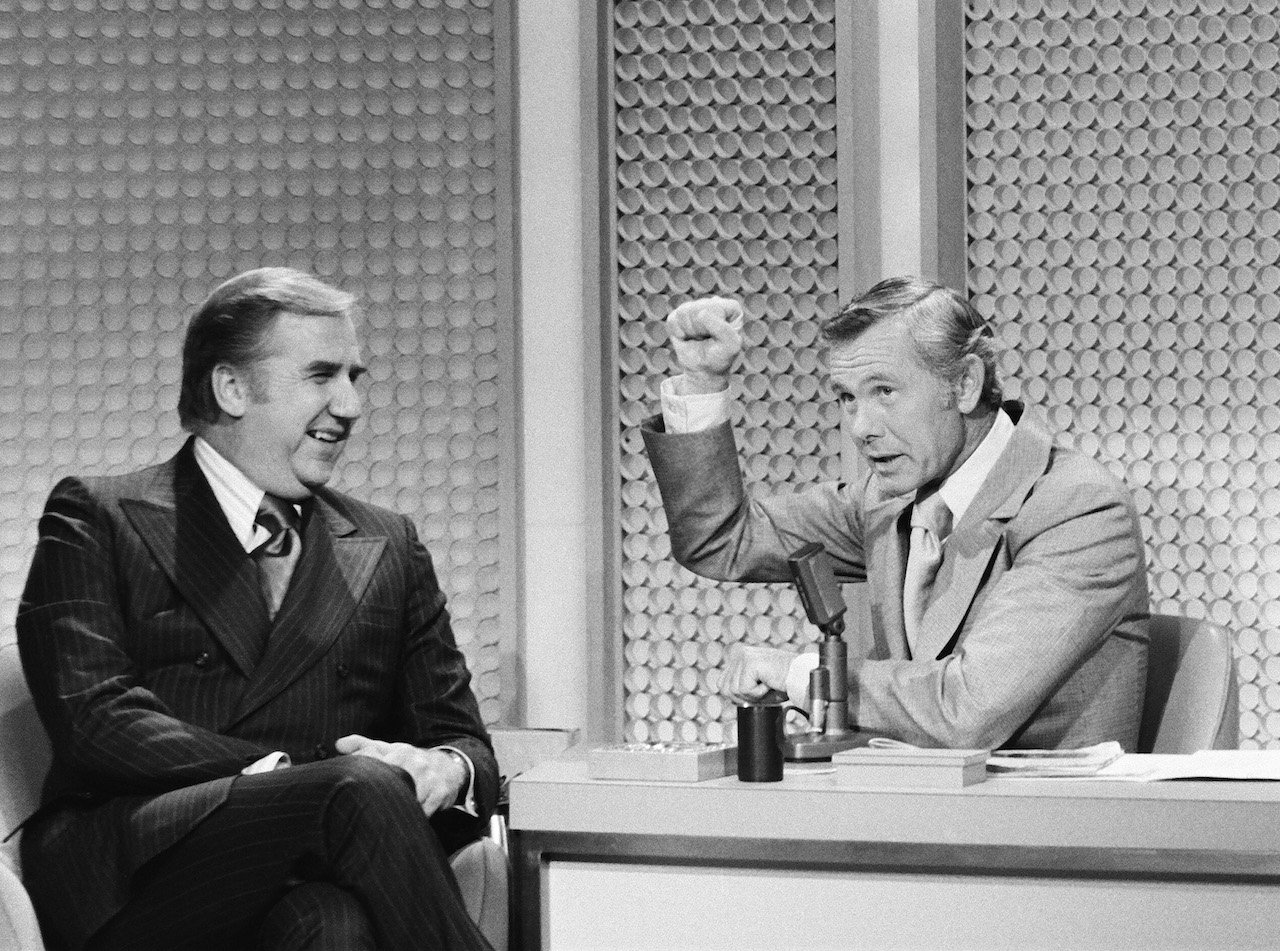 Ed McMahon sits next to Johnny Carson at The Tonight Show desk in 1972, as Carson raises one first in the air as if he's knocking
