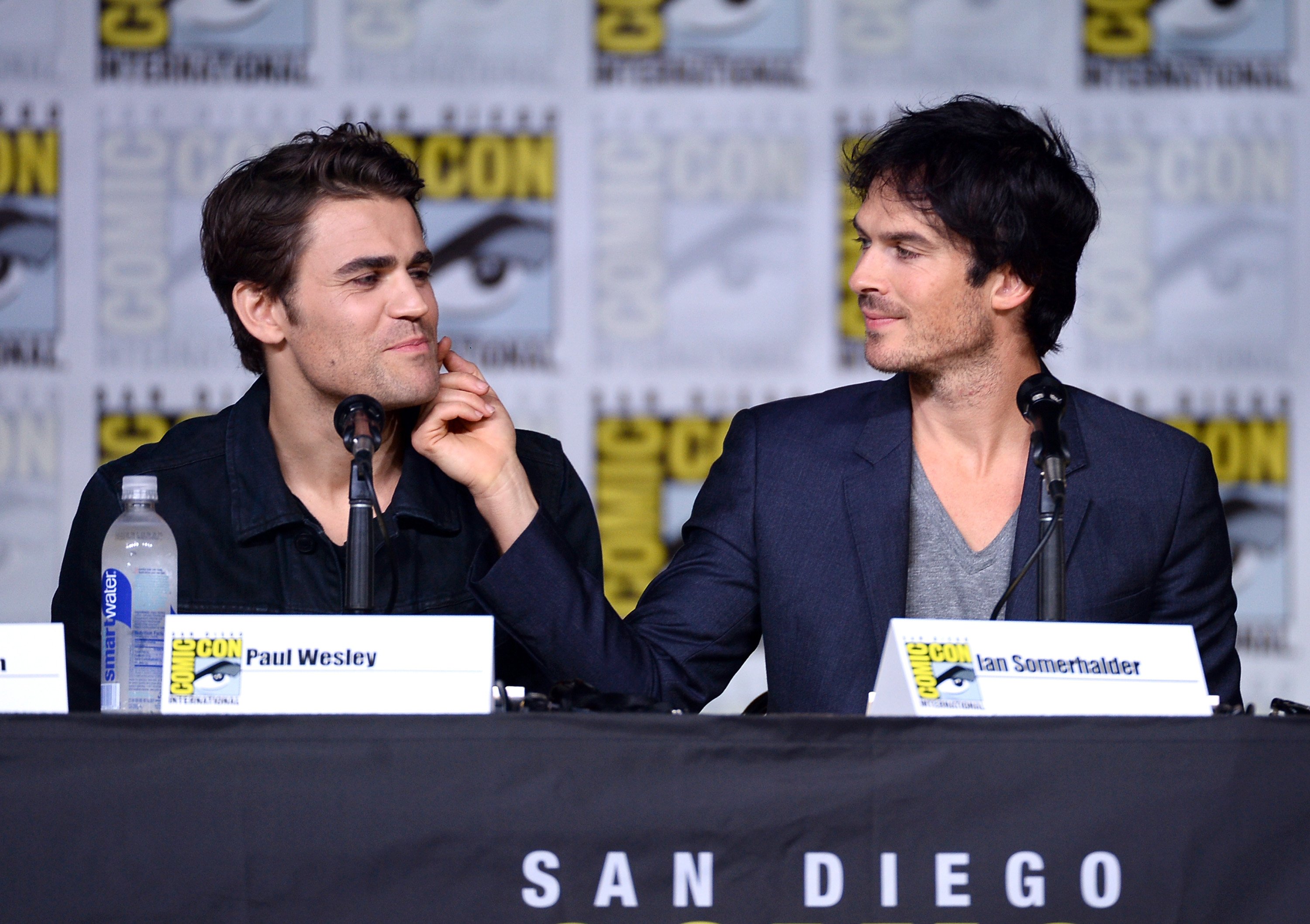 Paul Wesley and Ian Somerhalder, the cast of 'The Vampire Diaries,' sit onstage during a Comic-Con panel. Wesley wears a black button-up shirt. Somerhalder wears a blue suit over a gray shirt while brushing his hand across Wesley's cheek.