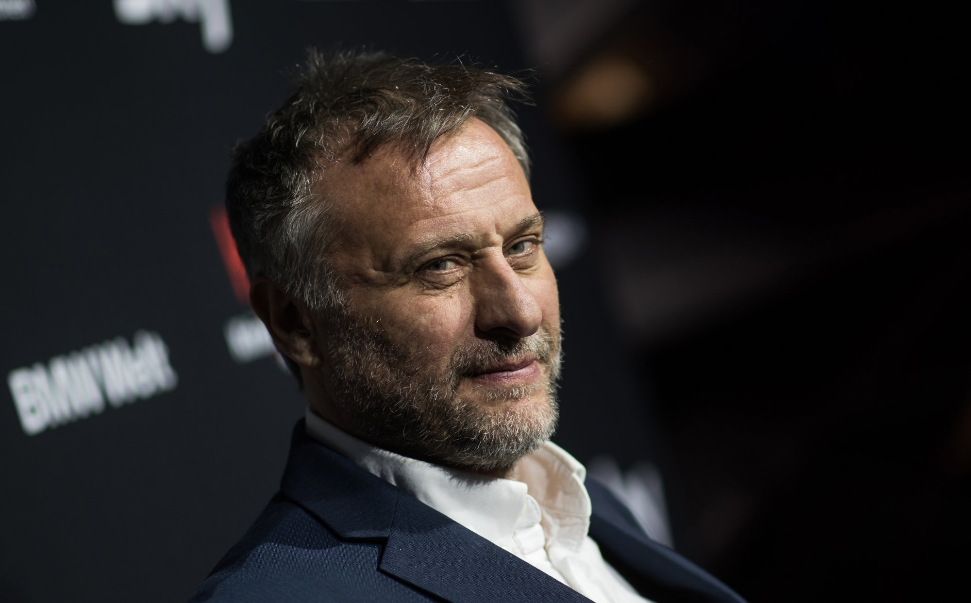 'John Wick' Michael Nyqvist wearing a navy blue suit jacket and a white dress shirt with a slight smile in front of step and repeat