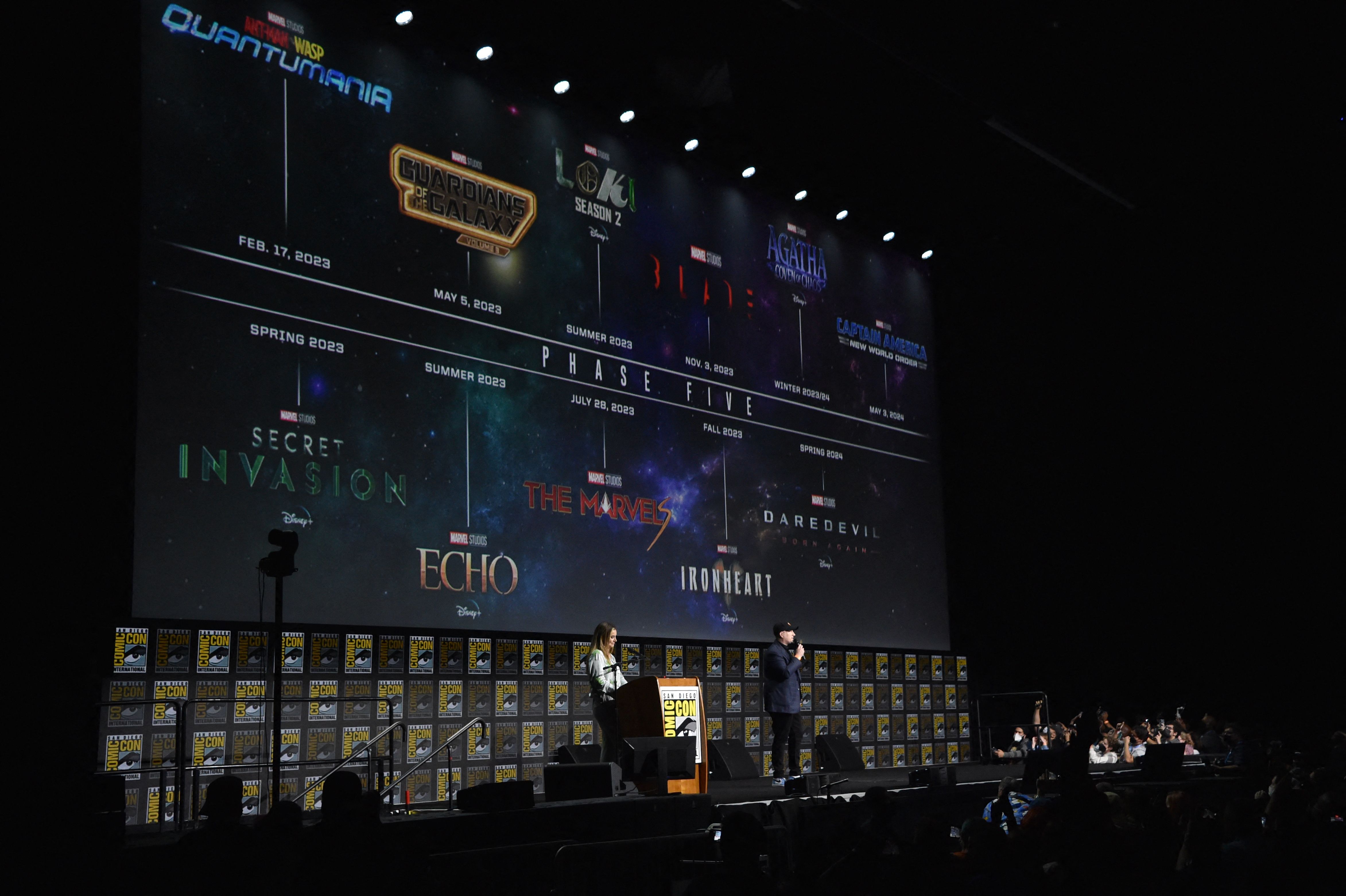 Kevin Feige appears onstage at San Diego Comic-Con 2022. He wears a gray suit, black pants, and a black baseball cap. Behind him is the MCU: Phase 5 schedule.