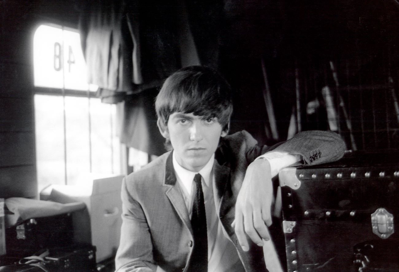 A black and white photo of George Harrison wearing a suit and resting his arm on a trunk.