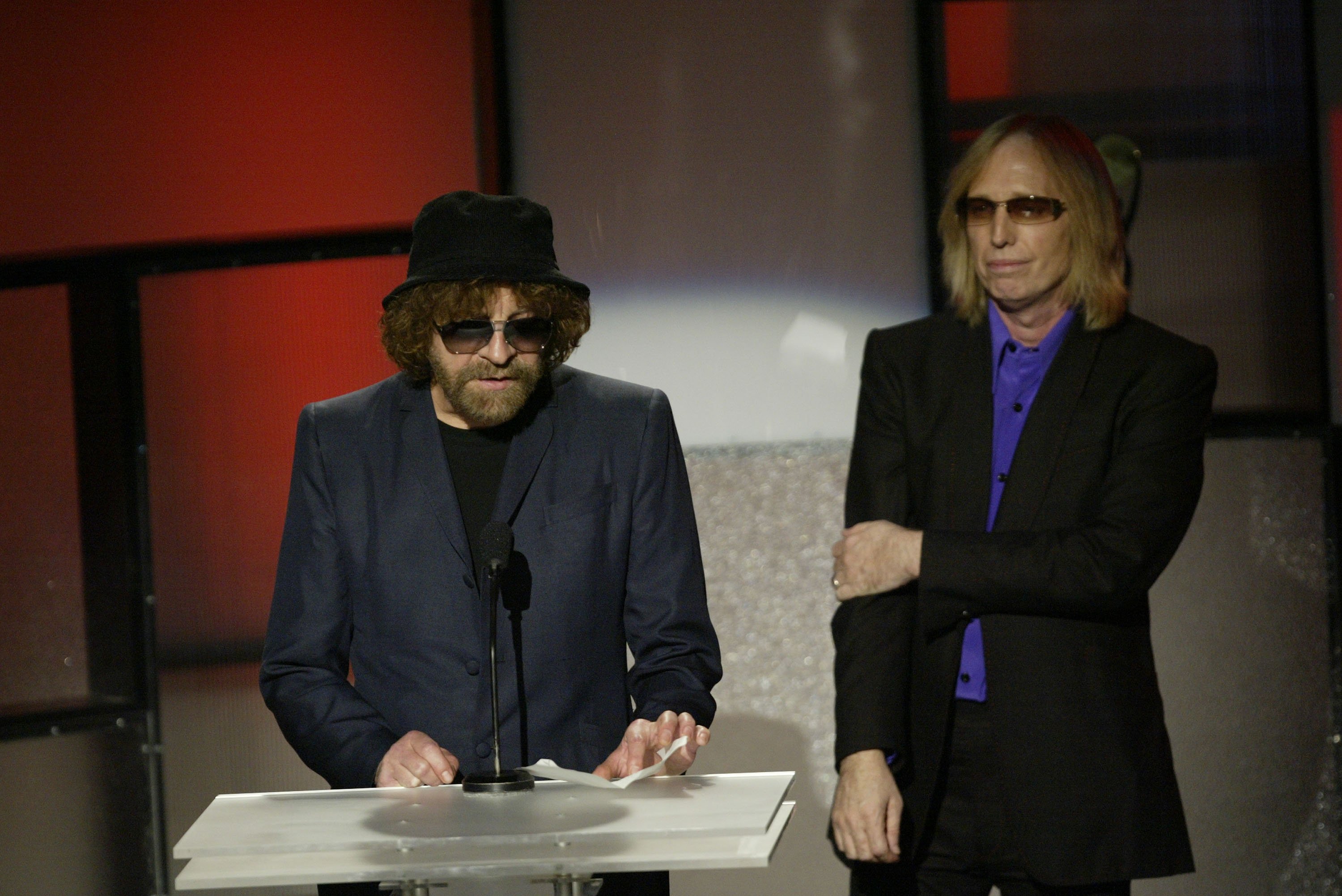 Jeff Lynne speaks into a microphone and Tom Petty stands next to him. Both wear sunglasses.