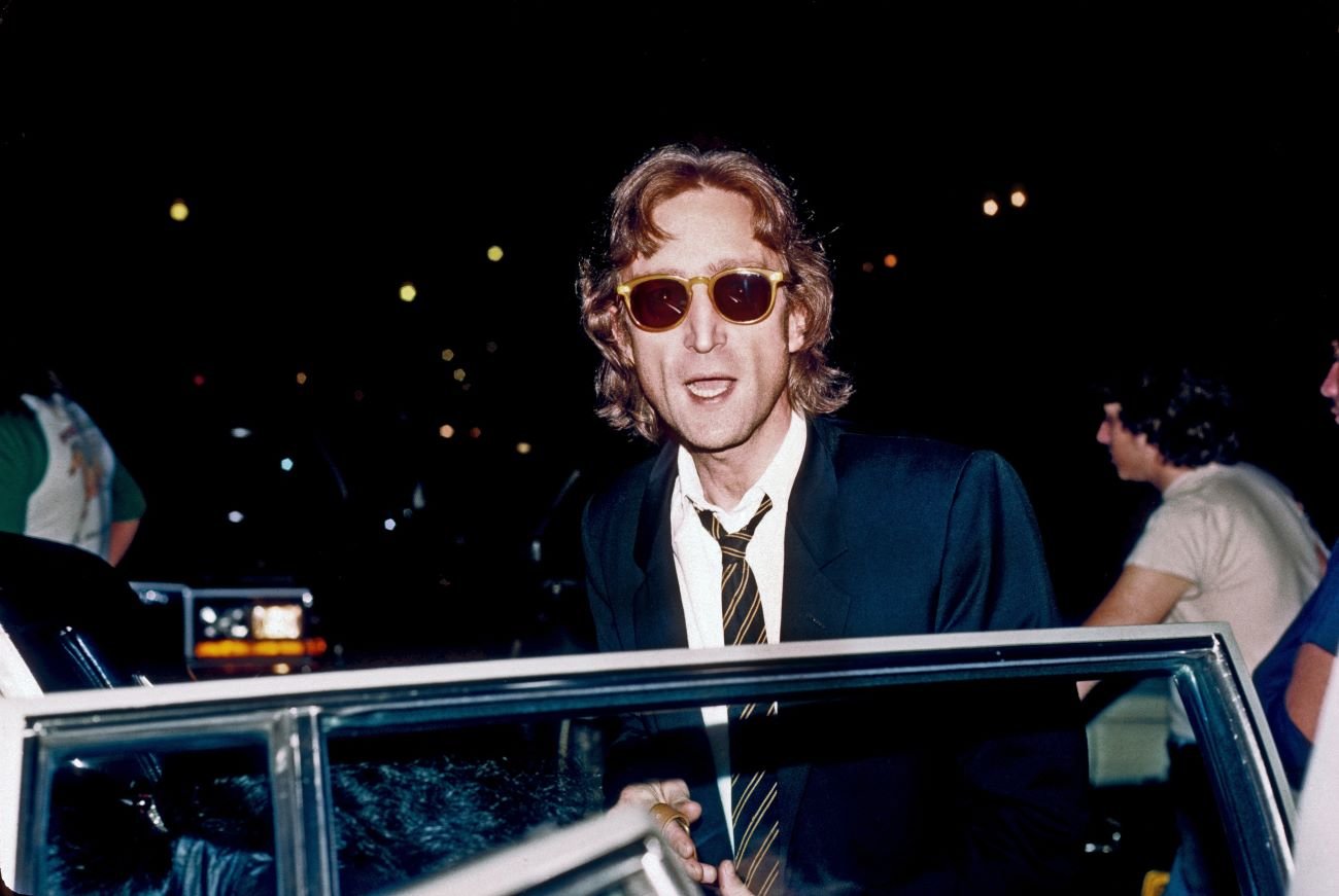 John Lennon wears sunglasses and a suit and stands outside a car.