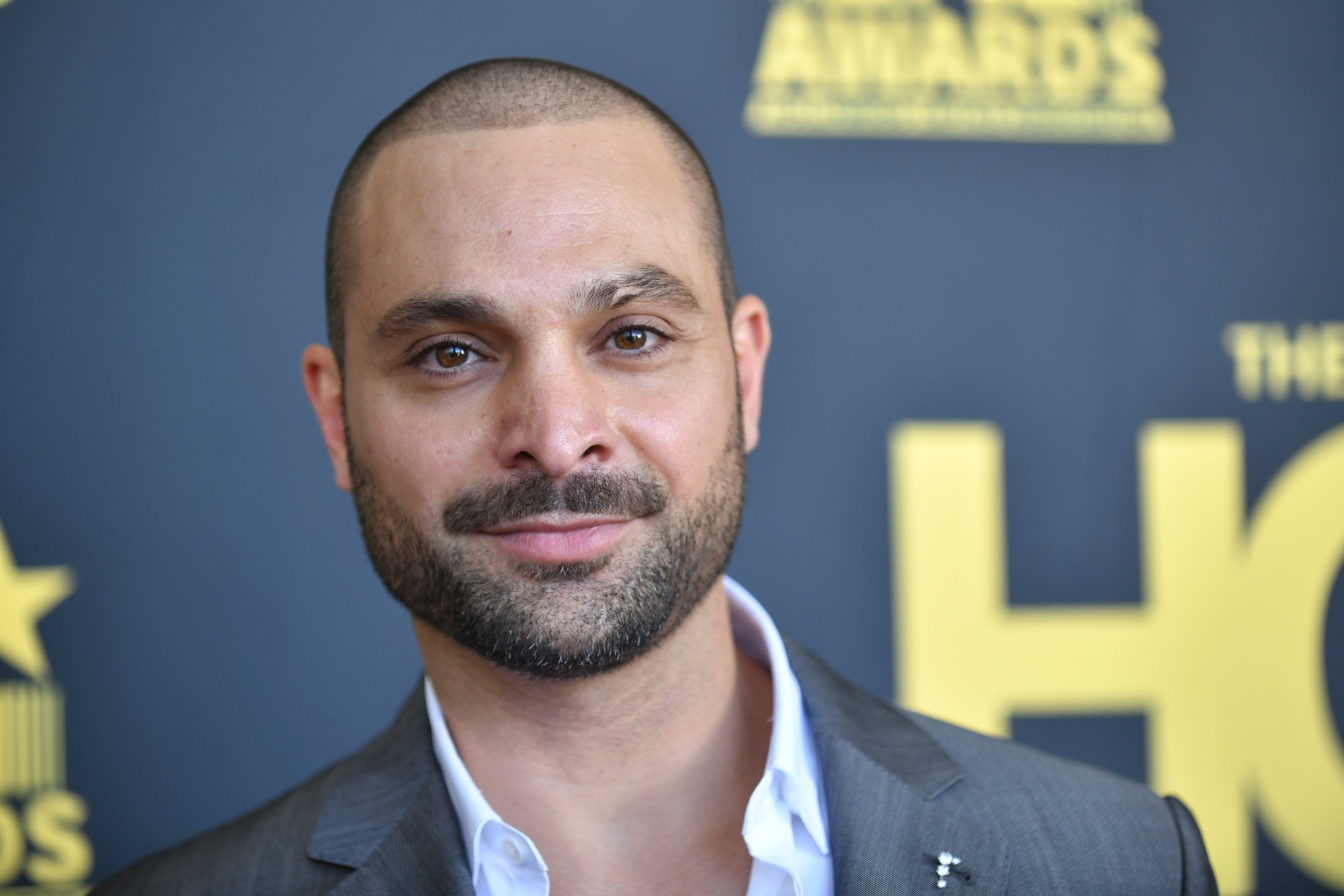 Michael Mando joins the 'Better Call Saul' cast at the the 2nd Annual HCA TV Awards - Broadcast & Cable. He's wearing a white shirt and grey jacket and smiling at the camera.