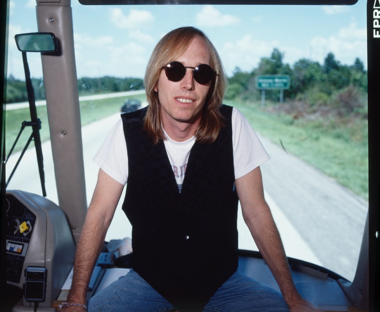 Tom Petty wears sunglasses and a vest and sits on a bus.