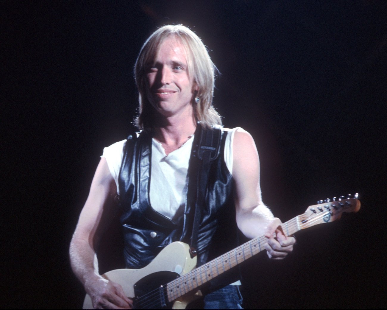 Tom Petty wears a black leather vest and holds a guitar.