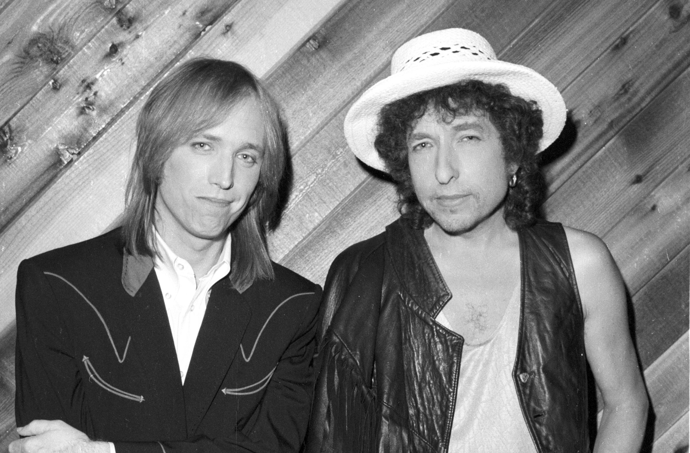 A black and white picture of Tom Petty and Bob Dylan posing in front of a wood paneled wall.