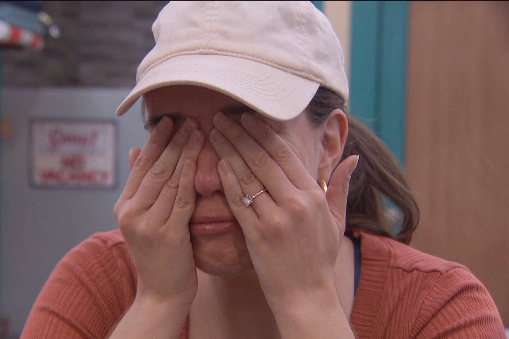 Brittany Hoopes, who, according to 'Big Brother 24' spoilers, is nominated during week 11, holds her hands over her eyes as she cries. Brittany wears a dark orange cardigan and light pink baseball cap.