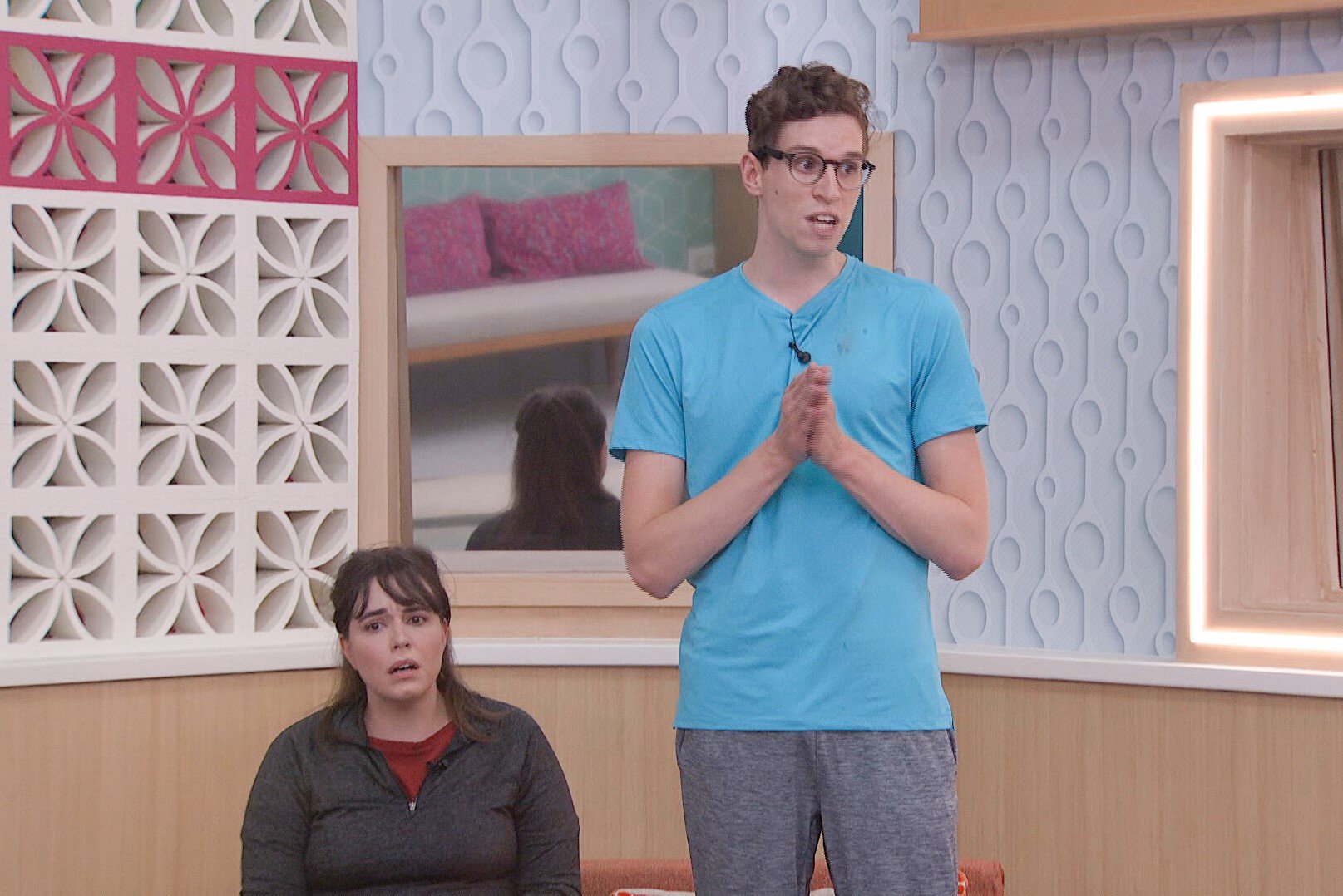 Brittany Hoopes and Michael Bruner during the 'Big Brother 24' double eviction. Brittany, who is sitting in the nomination chairs, wears a dark gray pullover over a red shirt. Michael, who is standing up to give a speech, wears a light blue shirt, gray pants, and glasses.