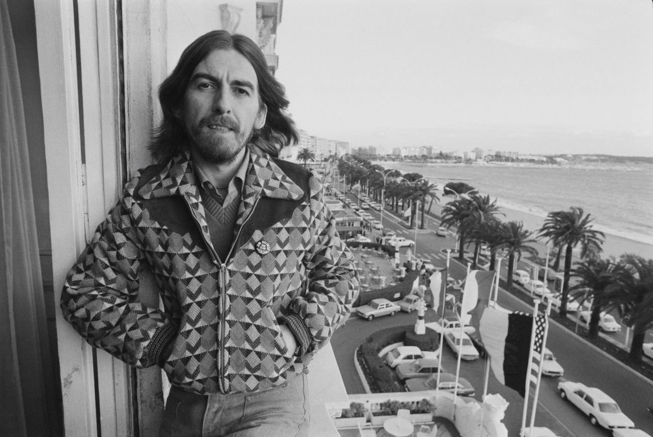 A black and white picture of George Harrison leaning against a window overlooking the beach.