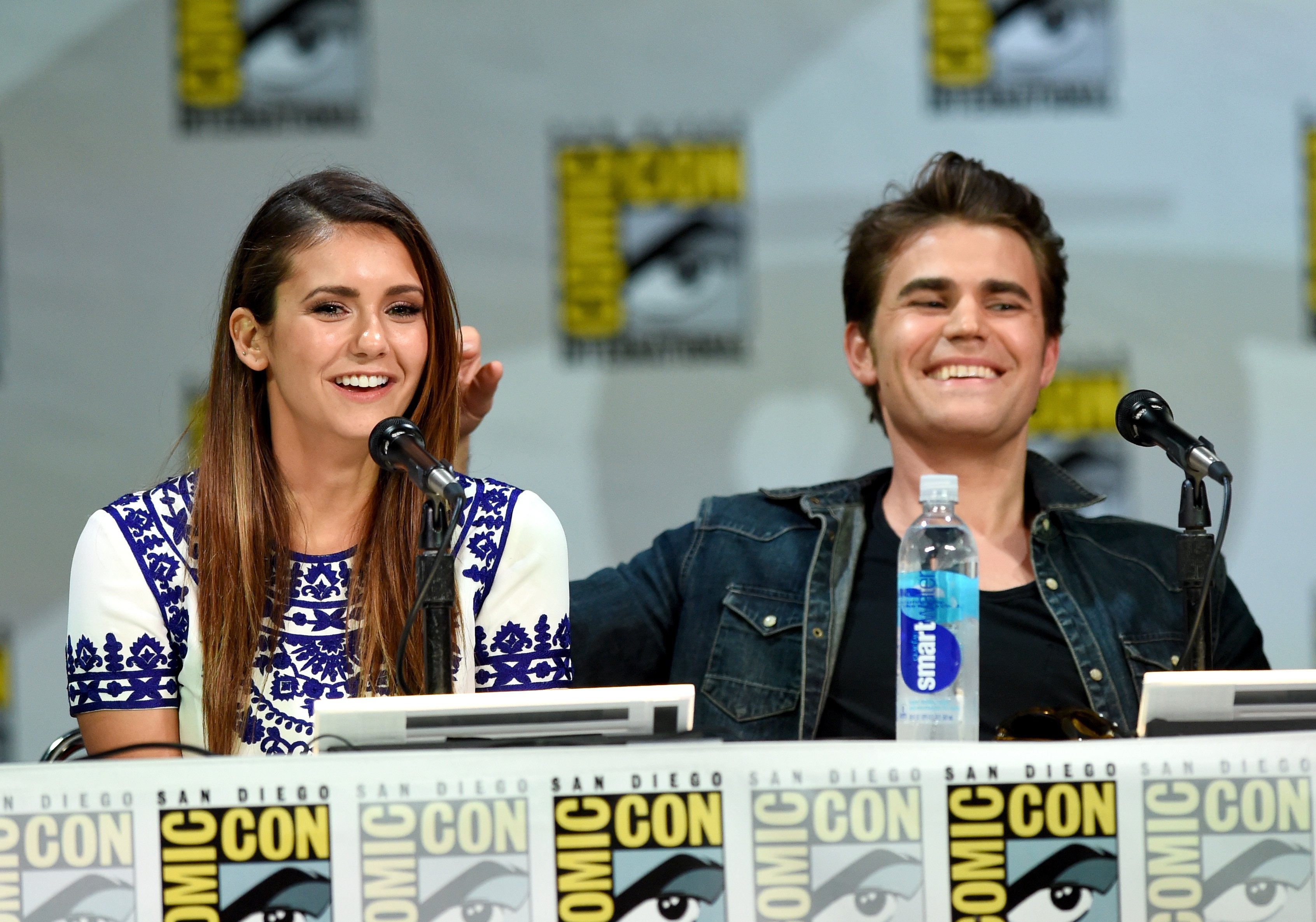 Nina Dobrev and Paul Wesley, whose show 'The Vampire Diaries' is leaving Netflix in September 2022, appear onstage at a San Diego Comic-Con panel. Dobrev wears a white and blue dress. Wesley wears a jean jacket over a black shirt.