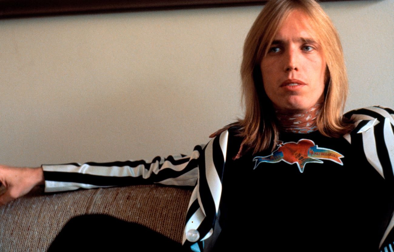 Tom Petty wears a black and white striped jacket and sits on a couch. 