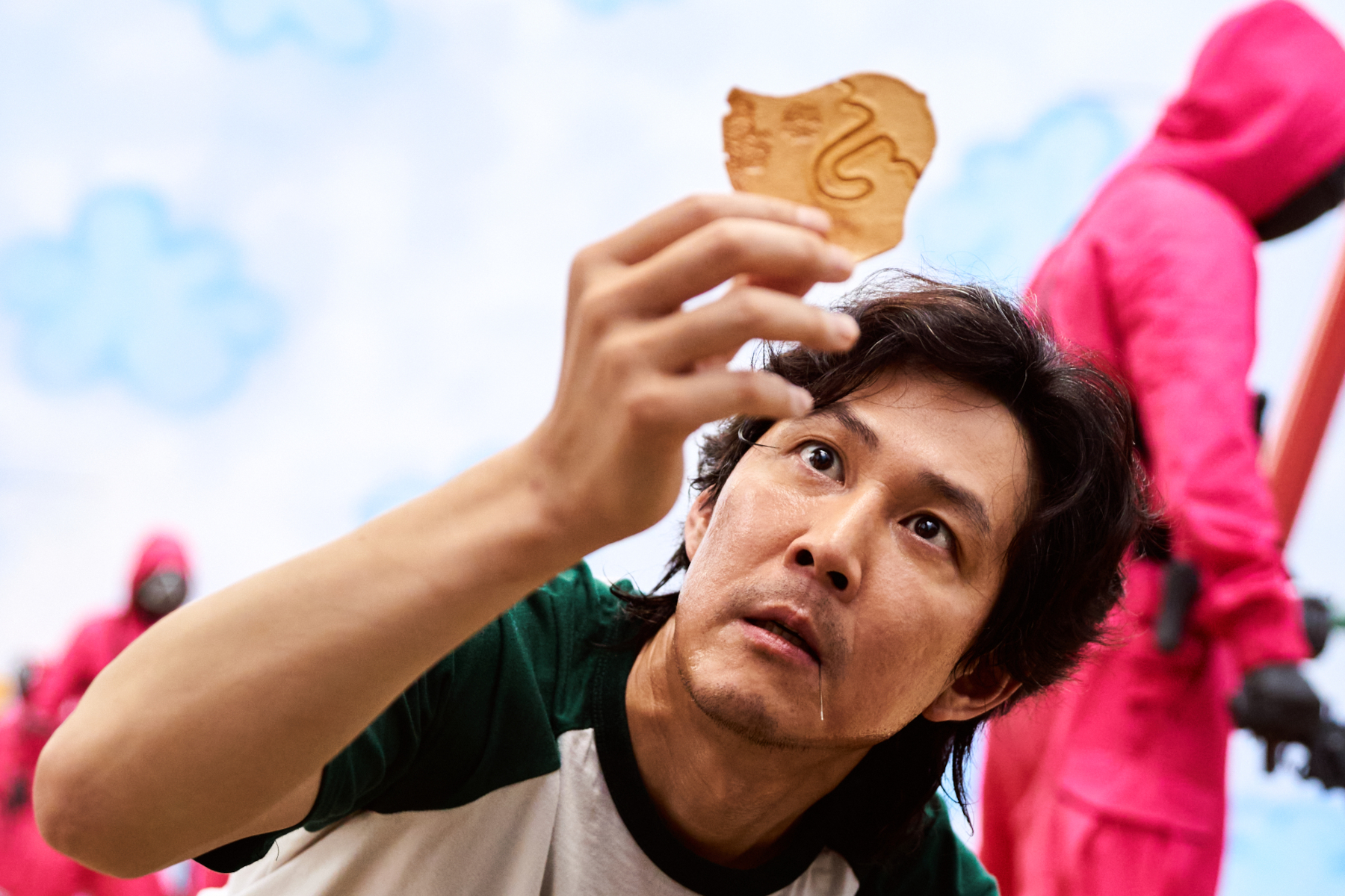 Lee Jung-jae as Seong Gi-hun in 'Squid Game' for our article about where to watch the series. He's wearing a green and white shirt and holding up a cracker with an umbrella carved into it.